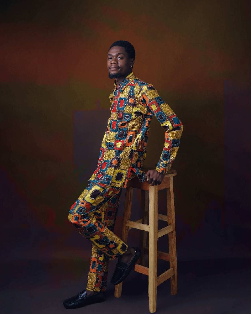 A man in a tailored patterned two-piece resting against a stool
